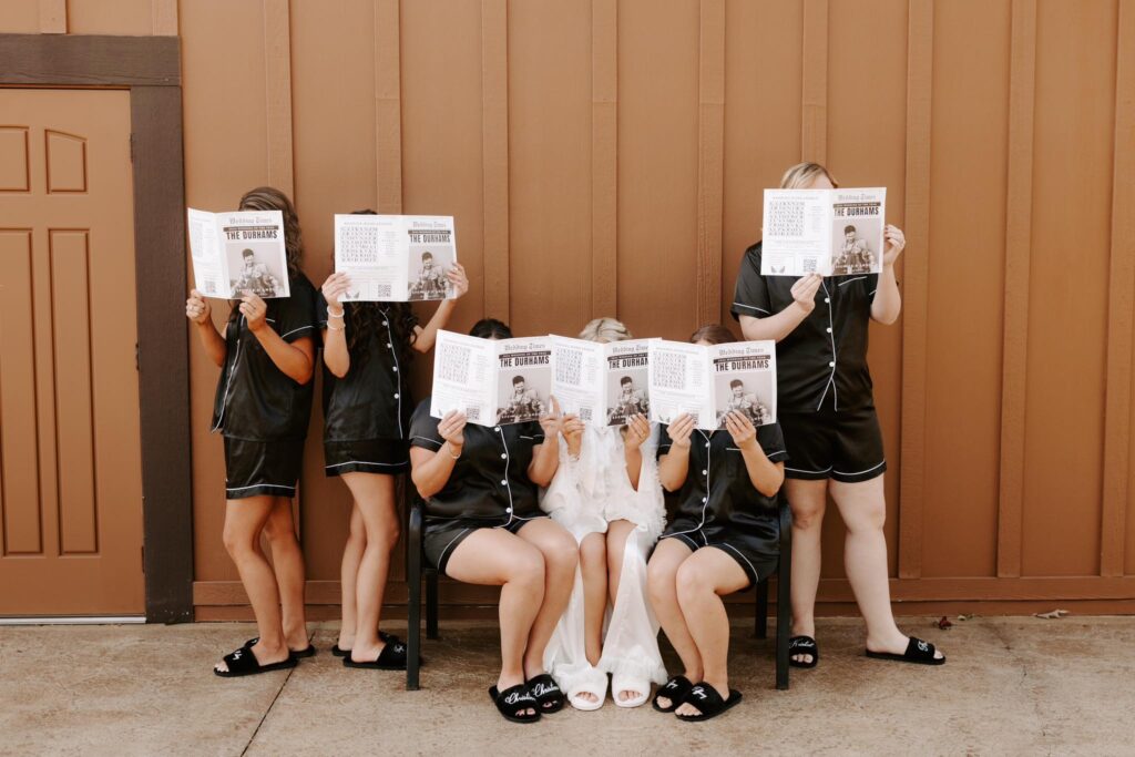Bridesmaids in matching black pajamas hold up personalized magazines while seated in front of a wooden backdrop, celebrating a wedding theme.