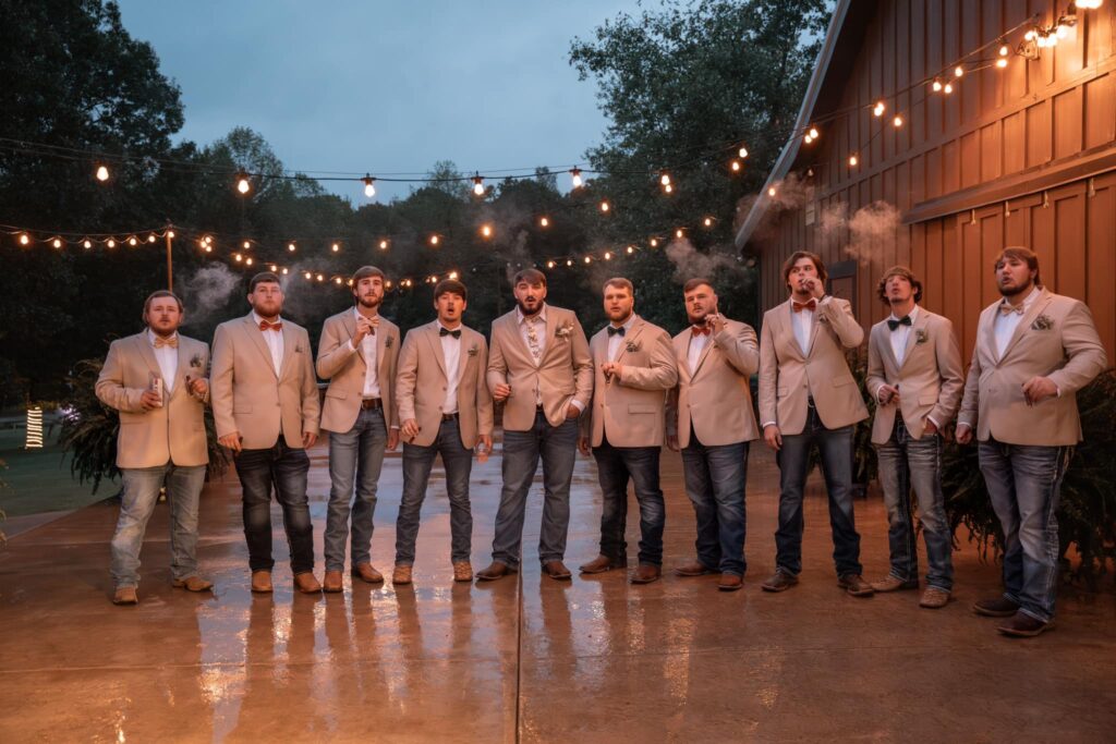 Groom and groomsmen in light suits pose outdoors under string lights, some holding cigars, creating a celebratory atmosphere.