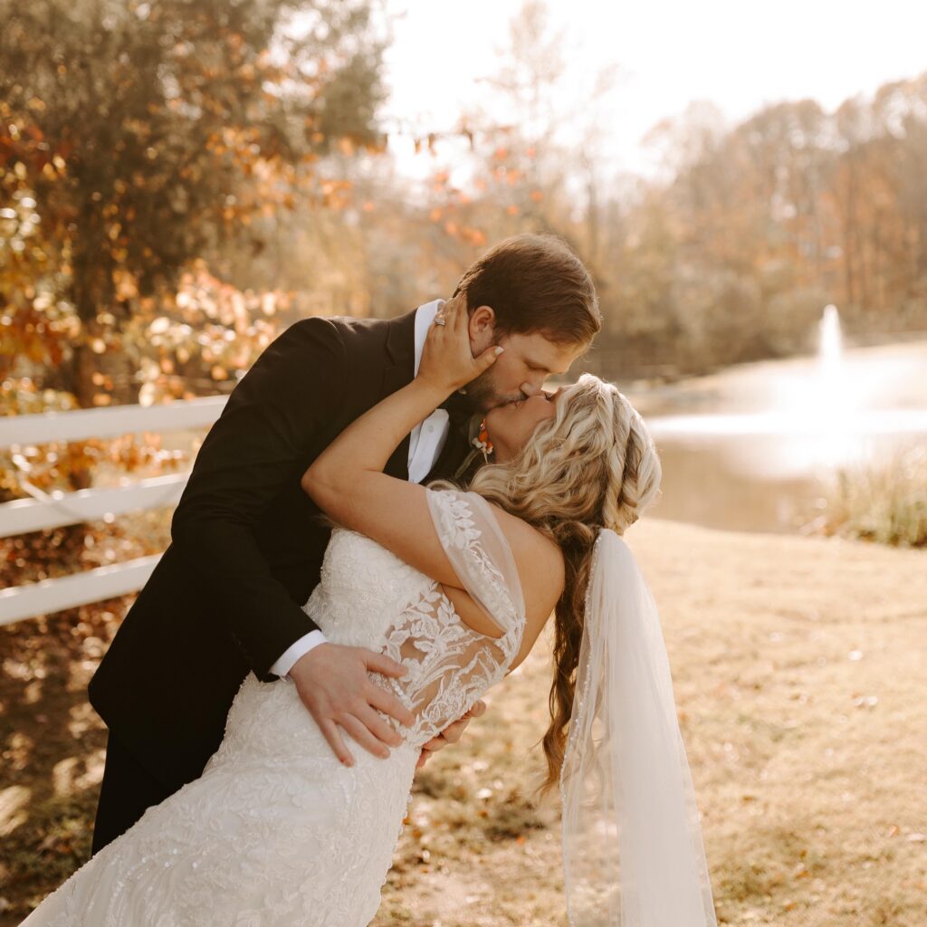 Bride in a lace gown and groom in a tuxedo share a romantic kiss outdoors, framed by autumn foliage and a serene pond in the background.