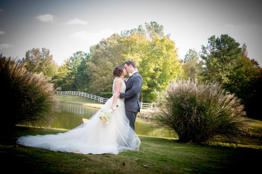 Bridal couple embracing near a serene pond, surrounded by lush trees and ornamental grass, capturing a romantic autumn wedding moment.