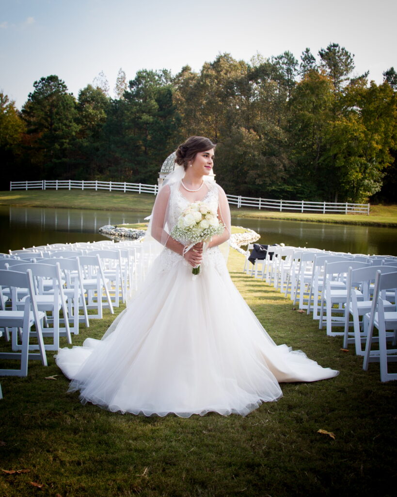 Bride in a flowing white gown holding a bouquet of white flowers, standing by a serene pond with rows of white chairs for a wedding ceremony.
