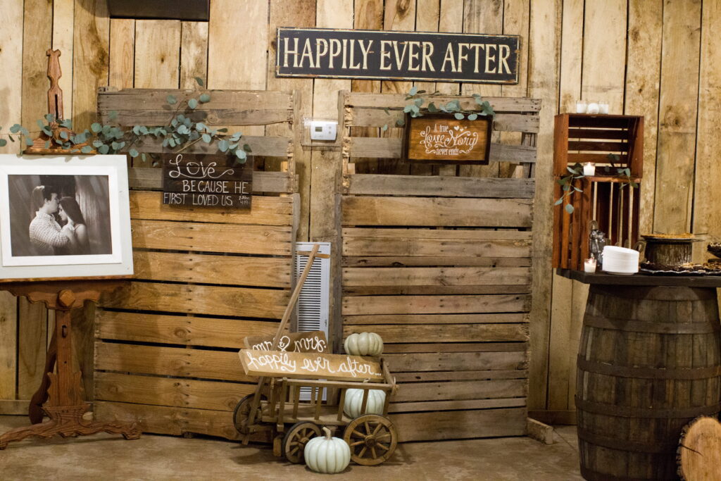 Rustic wedding decor featuring wooden backdrop, framed romantic photo, and signs proclaiming "Happily Ever After" and "Love because He first loved us."