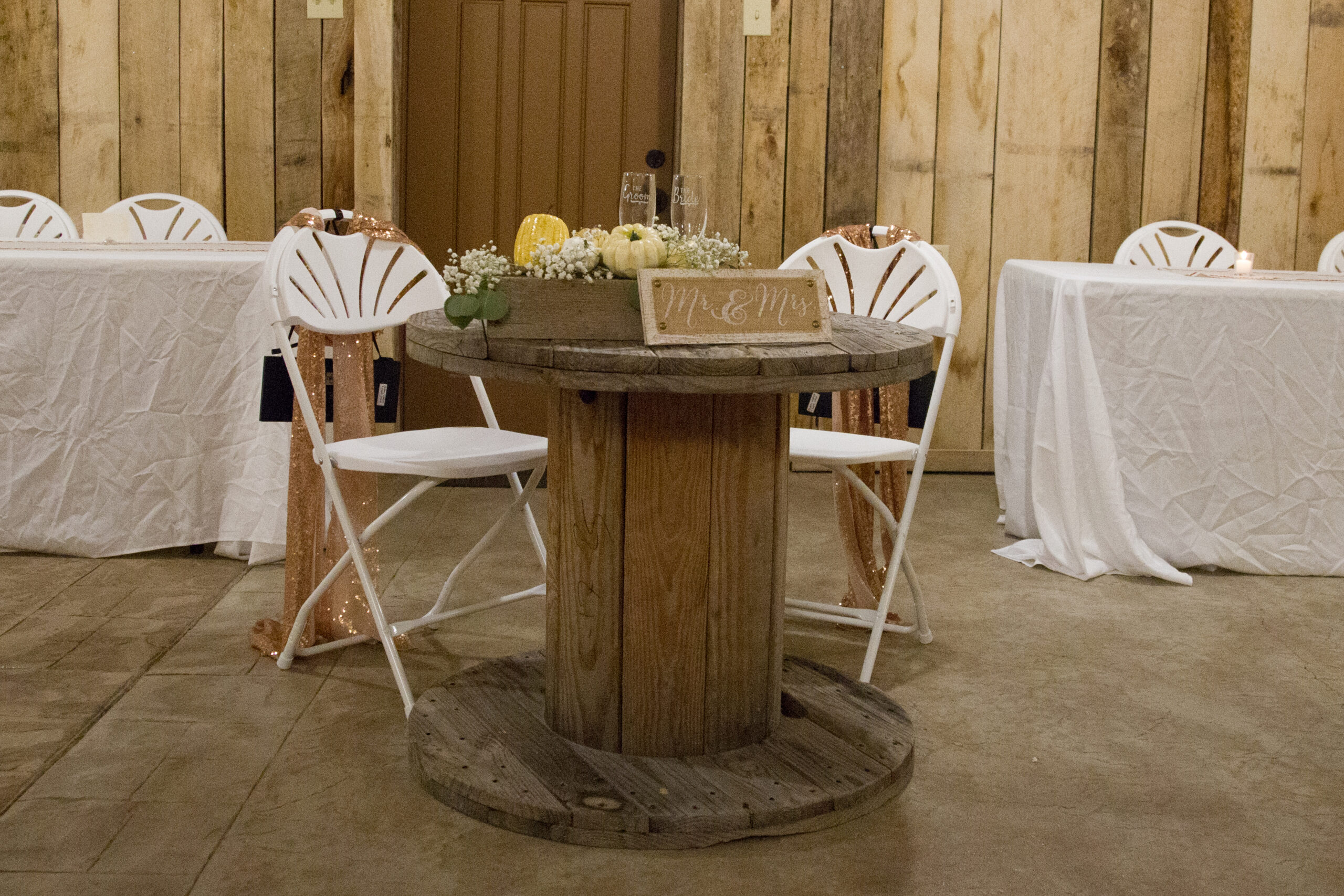 Rustic wedding decor featuring a round wooden table with a “Mr. & Mrs.” sign, paired with white chairs and simple floral arrangements.