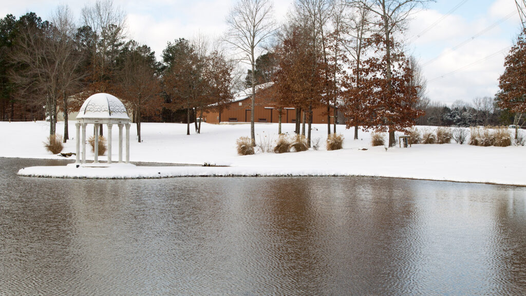 Snow-covered gazebo sits by a tranquil pond, with a backdrop of a rustic brown building and bare trees, capturing a serene winter scene.