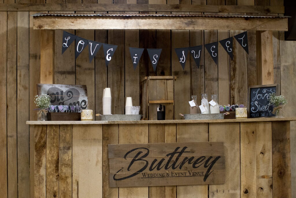 Rustic event bar at Buttrey Wedding & Event Venue, featuring a "Love is Sweet" banner, floral decor, and treats in decorated jars.
