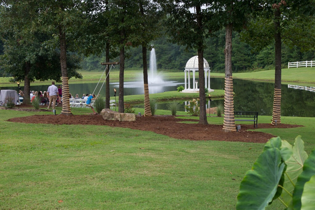 A serene outdoor venue featuring a pond with a fountain, surrounded by trees, a bench, and guests seated for an event near a decorative gazebo.