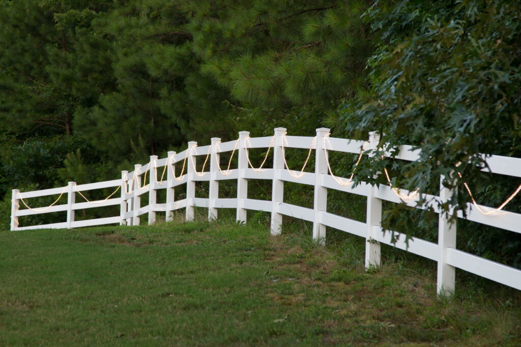 A white wooden fence adorned with warm string lights runs along a grassy area, bordered by trees, creating a serene outdoor ambiance.