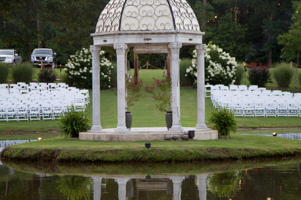 A scenic gazebo surrounded by a serene pond, with rows of white chairs set up for an outdoor event, likely a wedding ceremony.