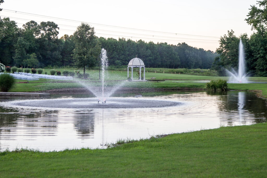 Serene landscape featuring two fountains in a pond surrounded by trees, with a gazebo and seating area in the background, ideal for events.