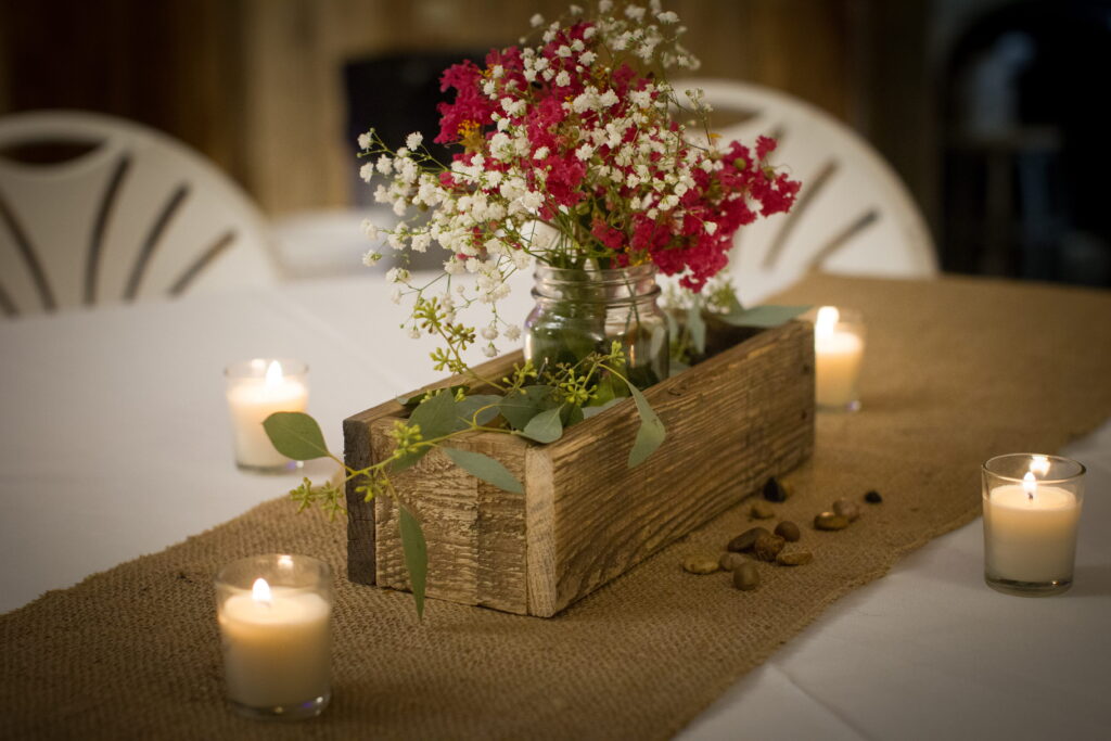 Rustic table centerpiece featuring a wooden planter with pink flowers, surrounded by candles on a burlap table runner, creating a cozy atmosphere.