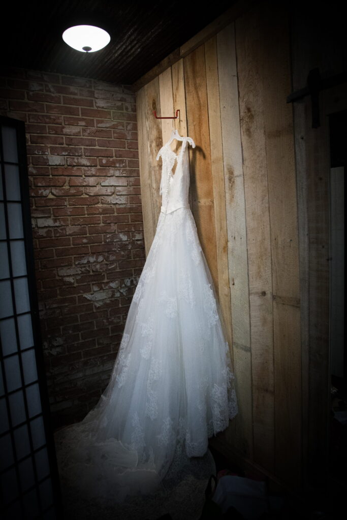 Elegant bridal gown hanging against a rustic wooden wall, illuminated softly. The image captures the anticipation of a wedding day.
