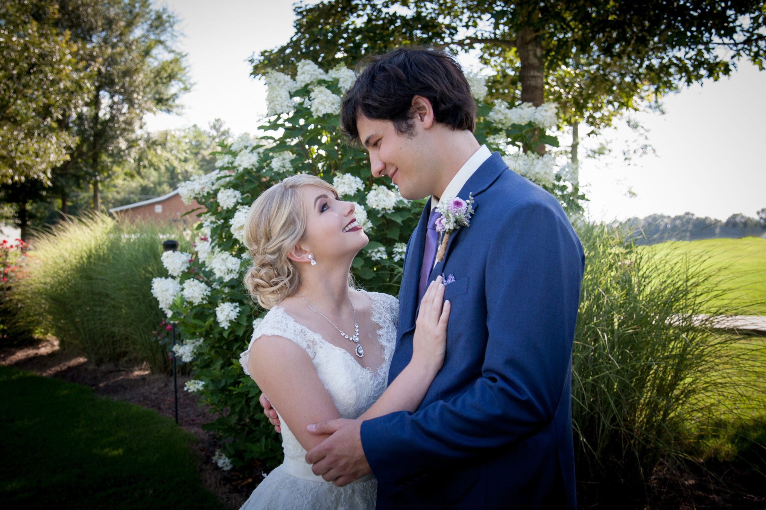 A bride in a lace gown and groom in a suit embrace amid lush greenery and white flowers, sharing a joyful moment on their wedding day.