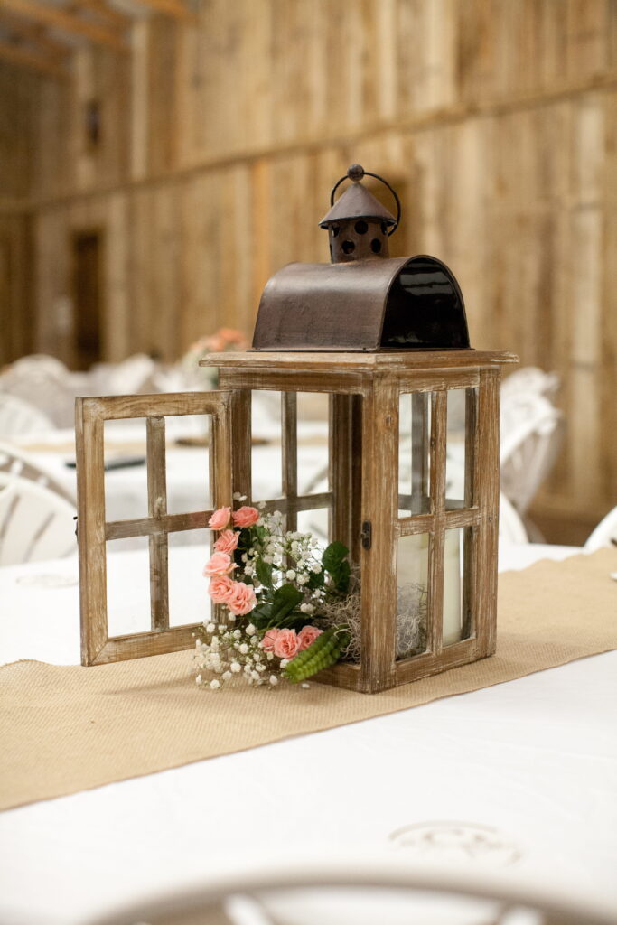Rustic lantern centerpiece features pink roses and greenery, set on a burlap table runner in a warm, understated venue.