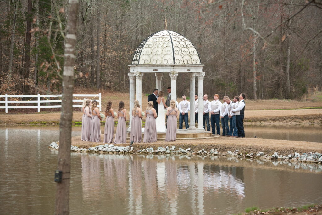 A wedding ceremony taking place by a serene pond, featuring a marble gazebo and a gathering of bridesmaids in blush dresses and groomsmen in white.