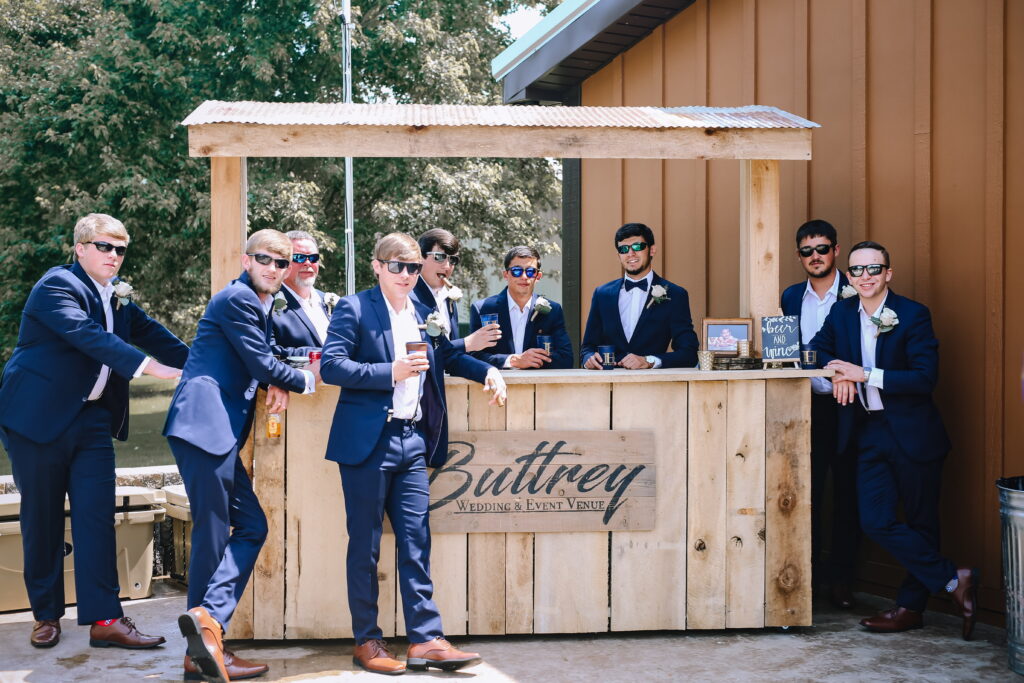 Groomsmen in navy suits and sunglasses gather around a rustic bar at Buttrey Wedding & Event Venue, enjoying drinks and celebration.