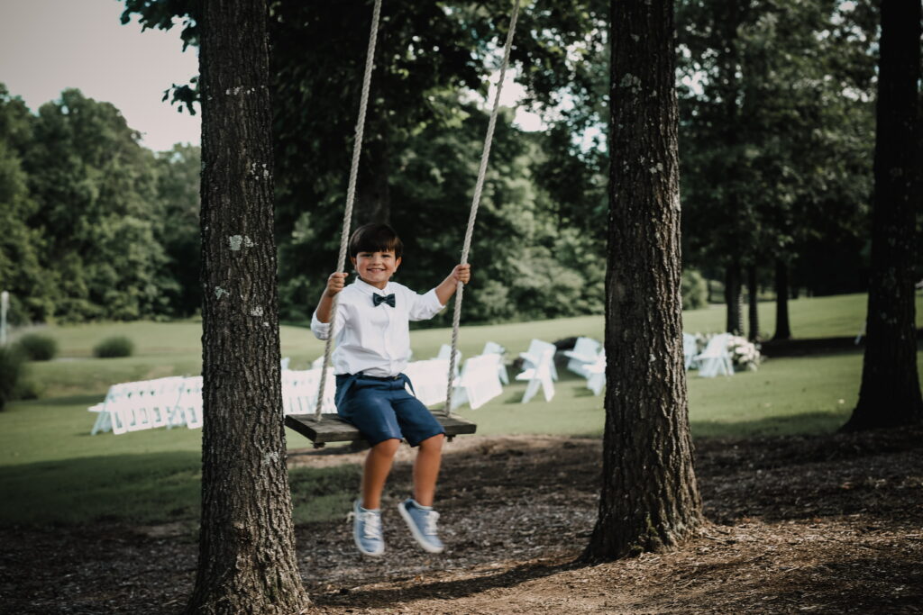 A young boy in a white shirt and bow tie happily swings between two trees, with a wedding setup visible in the background.