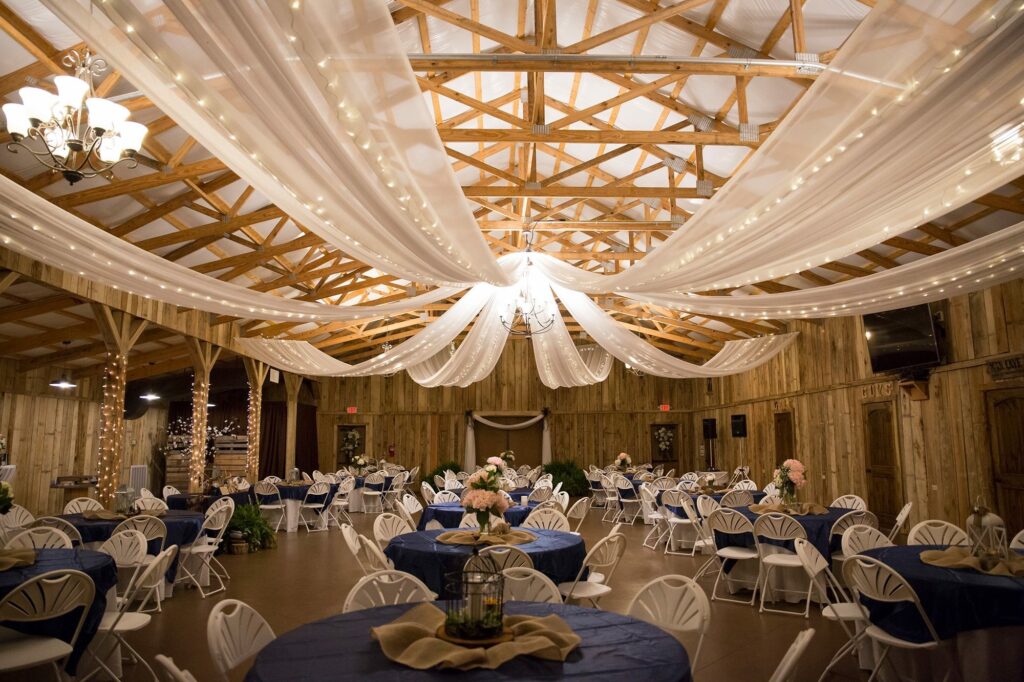 Elegant reception hall featuring draped fabric and string lights, set up with round tables and white chairs, ideal for events like weddings.