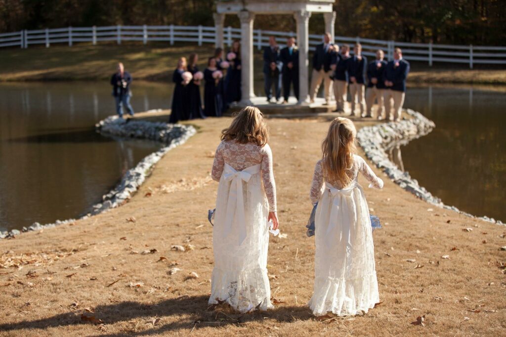 Two young girls in white dresses walk towards a wedding ceremony by a pond, with bridal party members in the background.