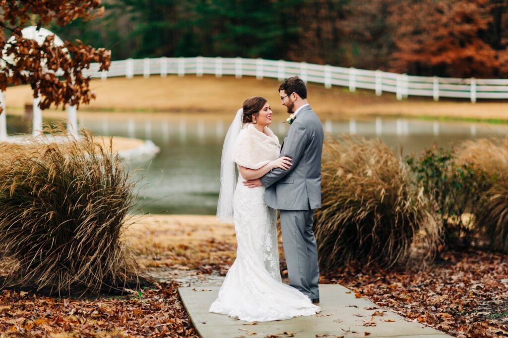 A bride and groom stand together by a serene pond, surrounded by tall grass and autumn foliage, sharing a tender moment.