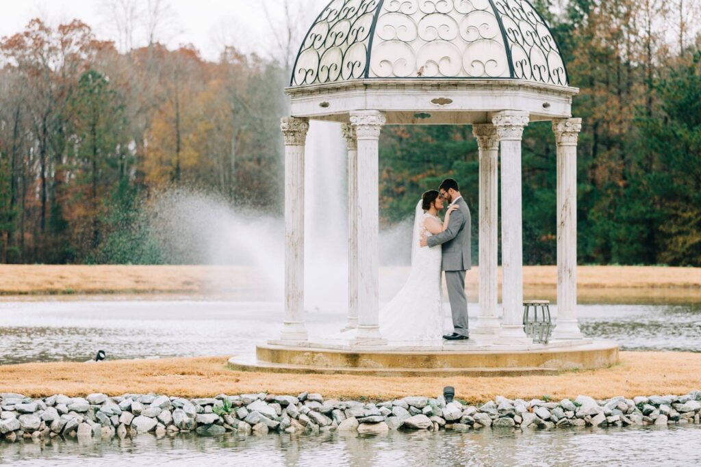 A bride and groom embrace under a decorative gazebo by a tranquil pond, capturing a romantic wedding moment amidst autumn foliage.
