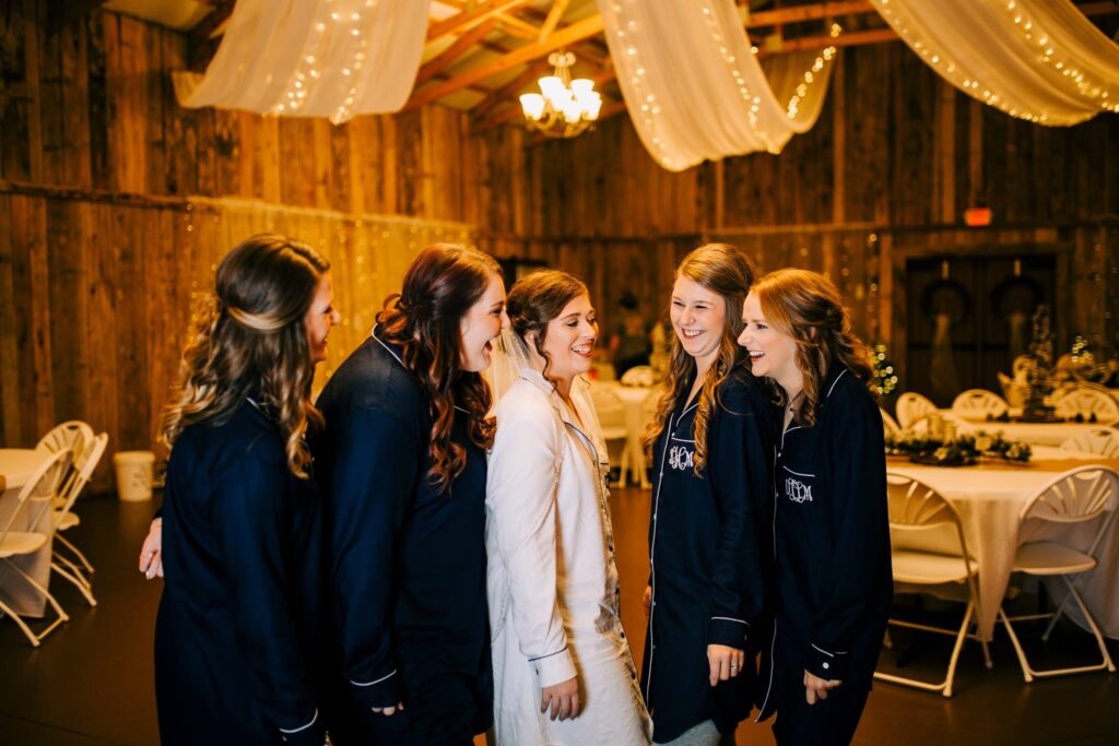 Bridesmaids in matching pajamas laugh with the bride in a rustic venue adorned with soft lights and elegant decorations, capturing a joyful moment before the ceremony.