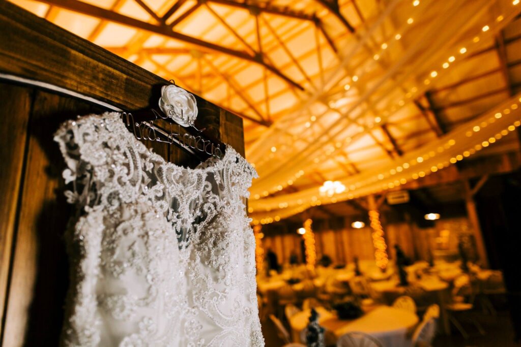 Delicate wedding dress adorned with intricate lace details hanging in a warmly lit barn venue, showcasing an elegant bridal preparation setting.
