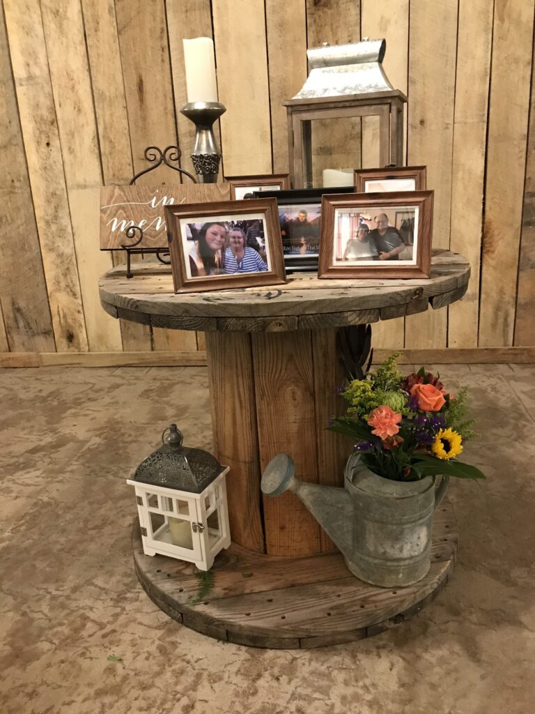 A rustic wooden table displays framed photos, a candle, and a watering can filled with flowers, set against a barn-like backdrop.