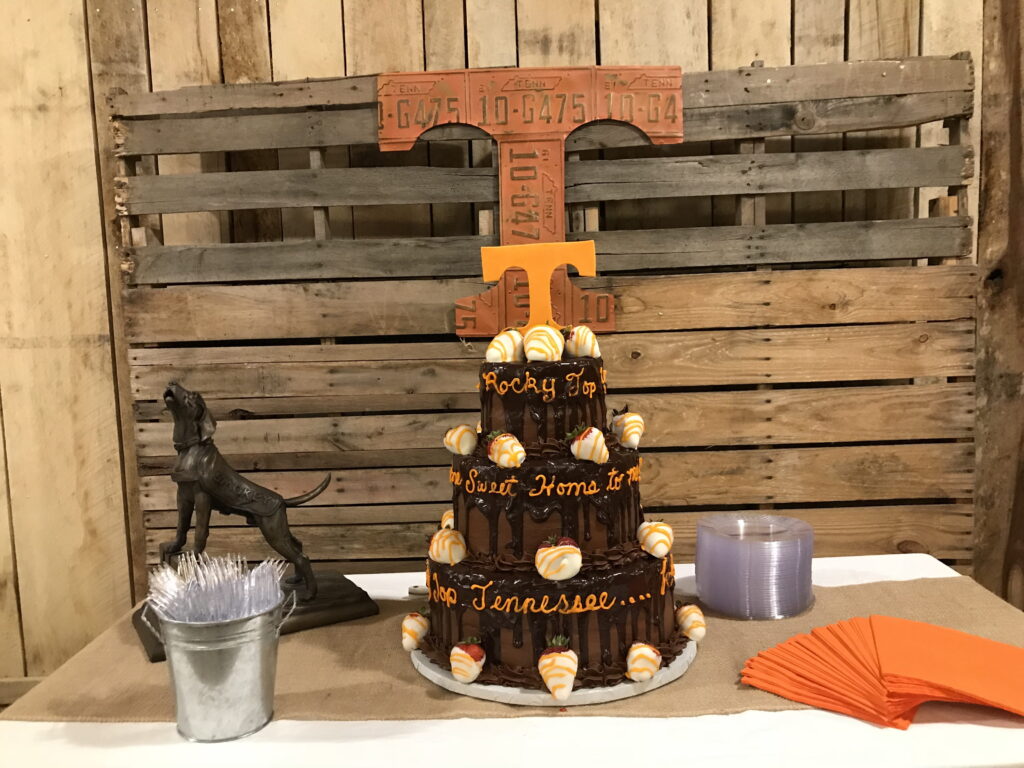 Tennessee-themed cake, shaped like a tower, features orange and white decorations with the phrase "Rocky Top, Sweet Home to Tennessee." A dog statue and party supplies are nearby.