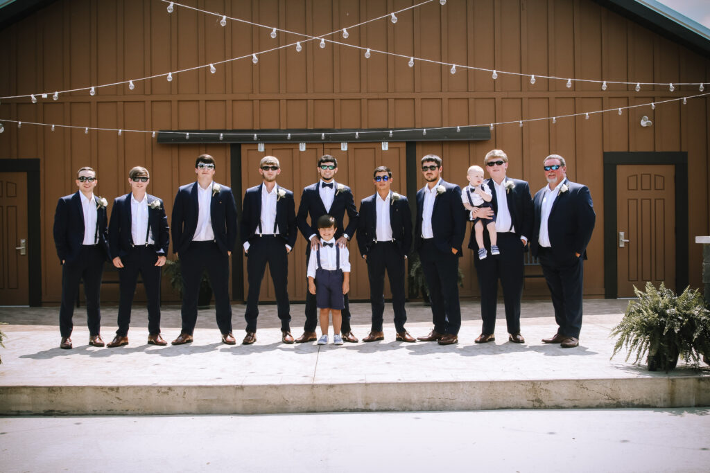 Groom and groomsmen, dressed in navy suits and sunglasses, pose on a sunny outdoor venue with a rustic backdrop, celebrating a wedding.