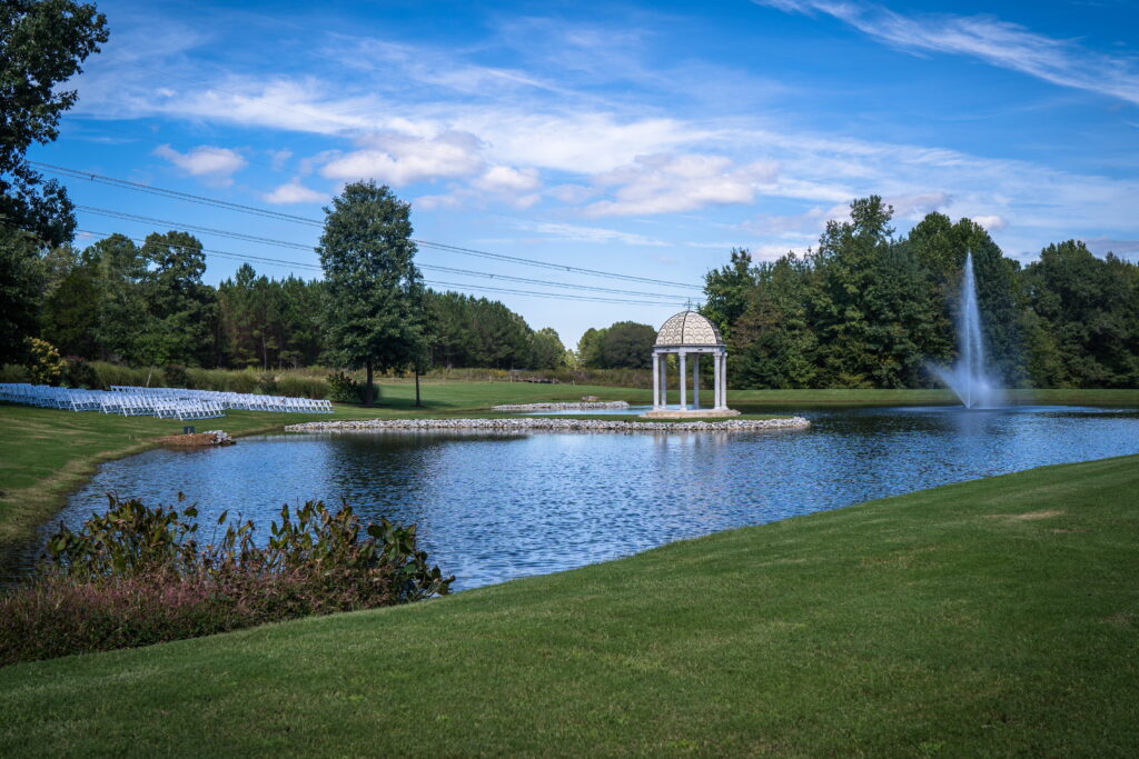 A serene outdoor venue featuring a pond with a decorative gazebo, surrounded by lush greenery and chairs arranged for an event.